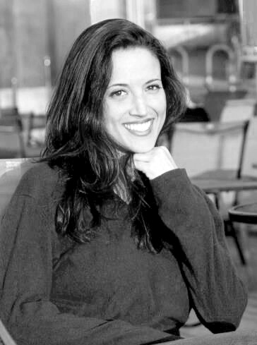 Ronna Bradus looking straight ahead and smiling, with one hand resting under her chin. She has long, dark hair and is wearing a dark-colored shirt. Black and white portrait.
