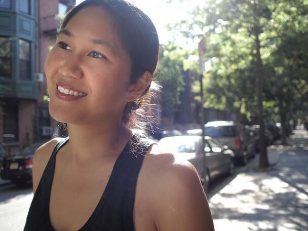 Stephanie Wang-Breal smiles, looking away from the camera. She is wearing a black tank top and standing on a sidewalk with trees, cars, a building, and the sun shining behind her.
