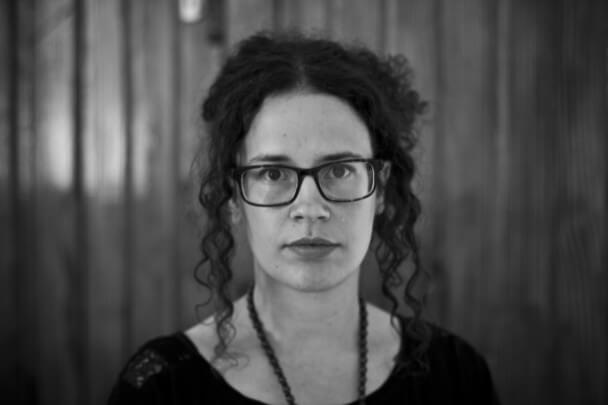 Naziha Arebi looking directly ahead. She is wearing dark-rimmed glasses and her curly hair is tied up with some tendrils loose on both sides of her face. Black and white portrait, with an out of focus background.