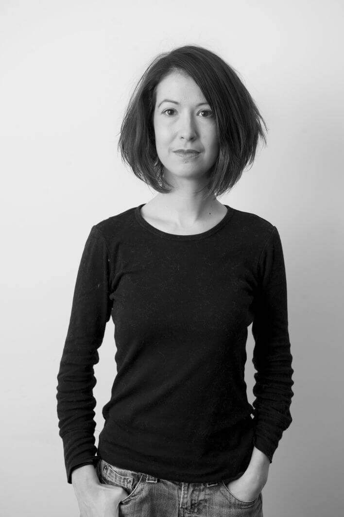 Francesca Panetta looking directly at the camera, standing in front of a white background. She has medium-dark hair, a dark long-sleeve t-shirt, and denim jeans. Portrait in black and white.