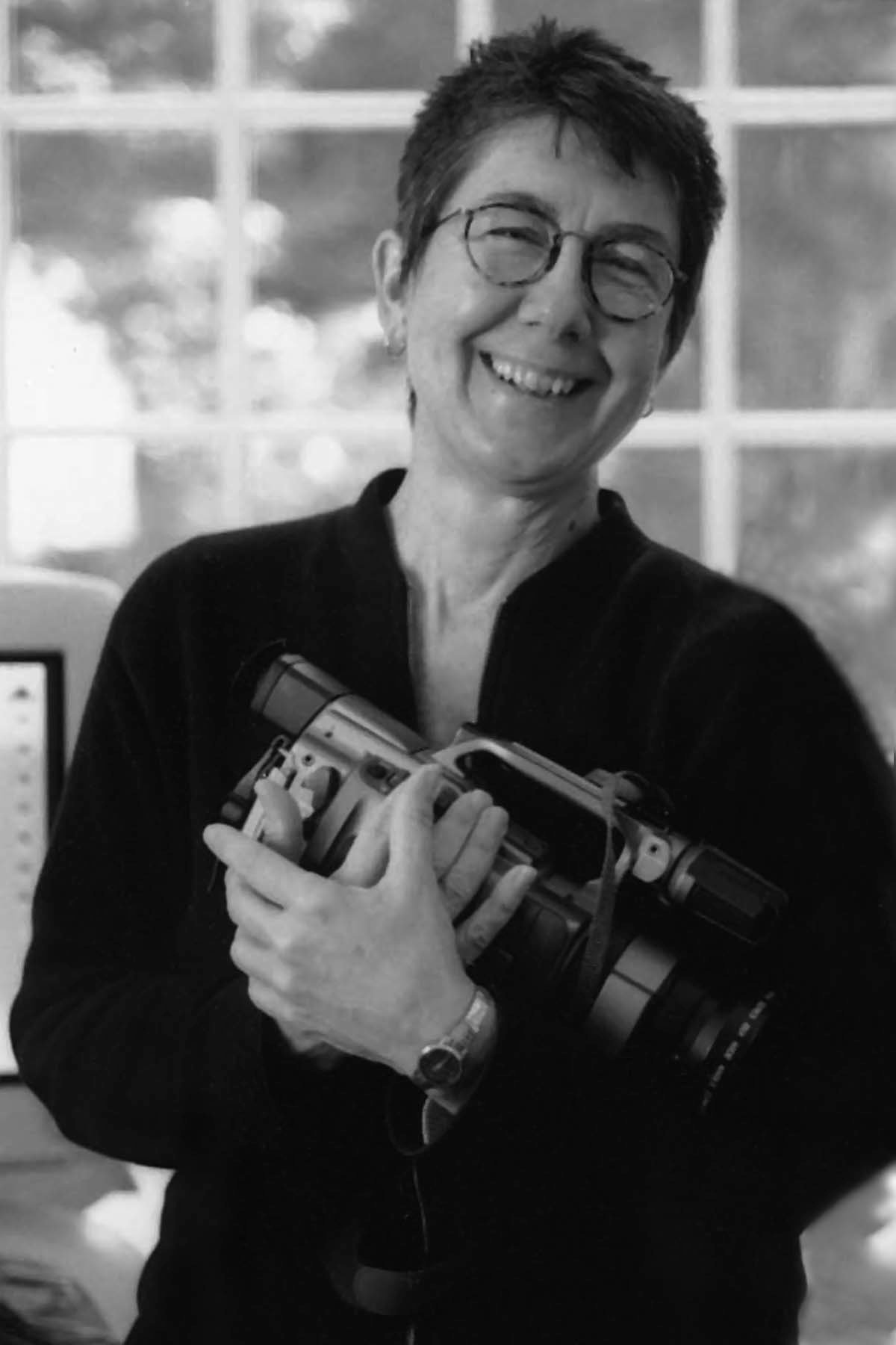 Julia Reichert wearing a dark blouse and holding a camera in her arms. She is smiling, has short hair and circled frames. Black and white portrait.