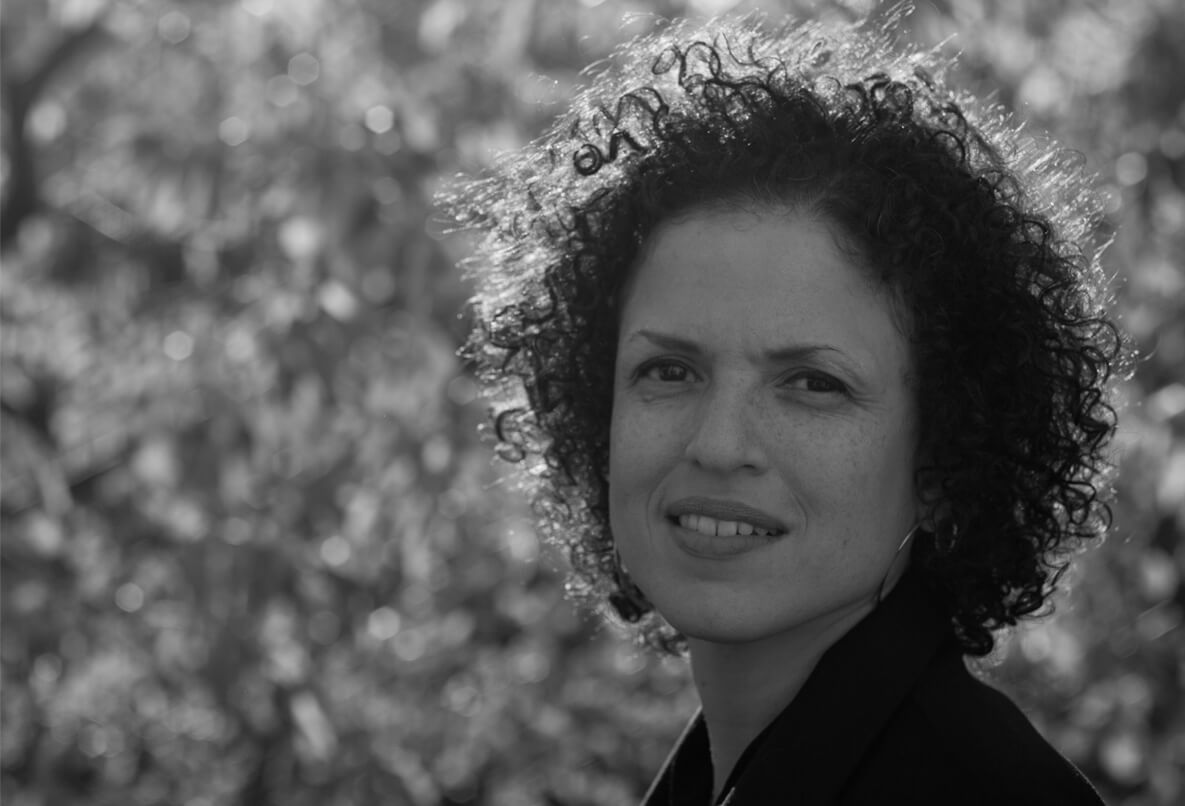 Michèle Stephenson looks at the camera, has curly short hair, and wears hoop earrings. Black and white portrait, trees out of focus in the background.