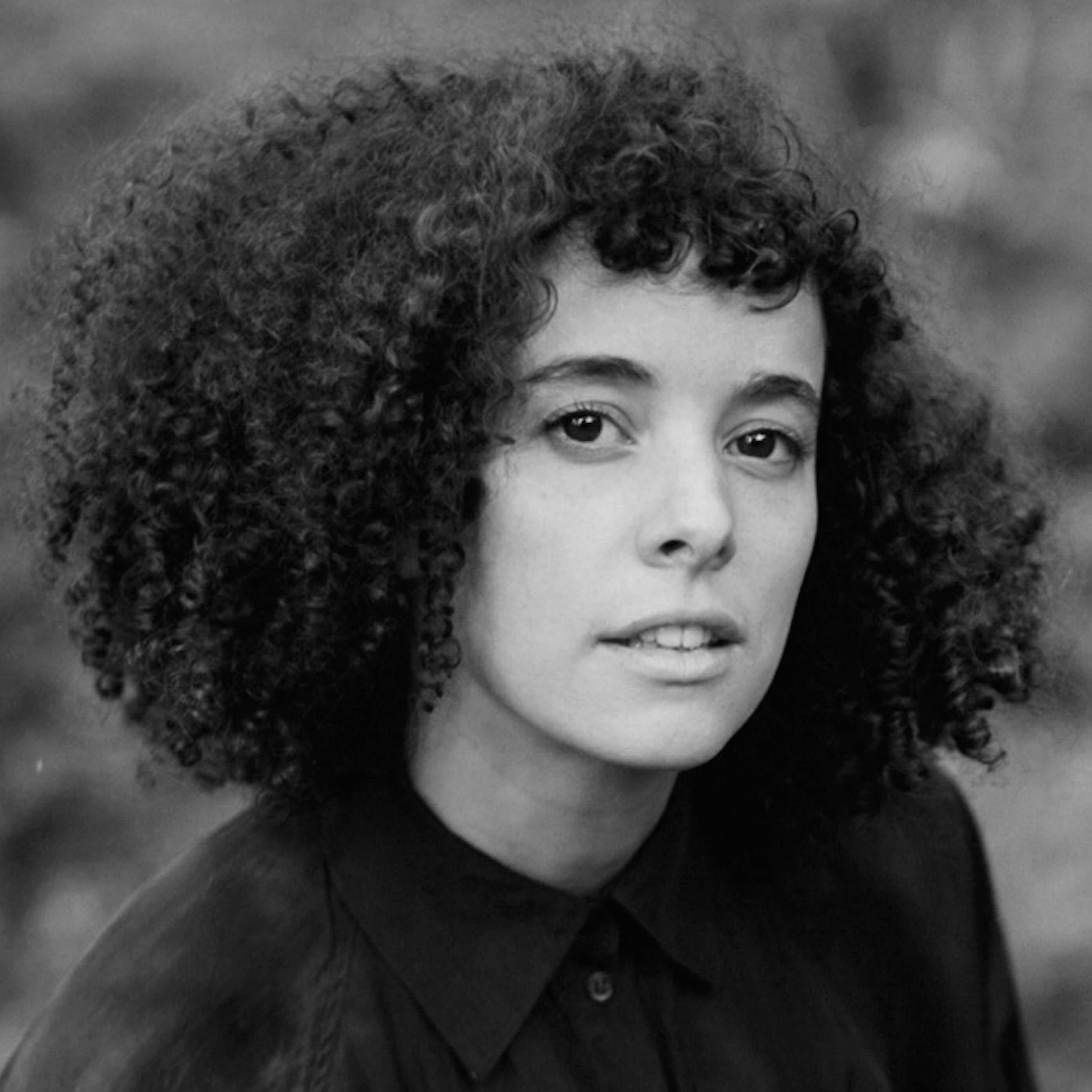 Malika Zouhali-Worrall looking straight ahead. She has dark, curly, shoulder-length hair and short bangs. Black and white portrait, with out of focus background.