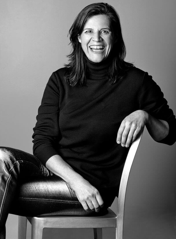 Kirsten Johnson siitting in a chair, wearing metallic pants and a turtleneck shirt. She has medium hair and is widely smiling at the camera. Black and white portrait.