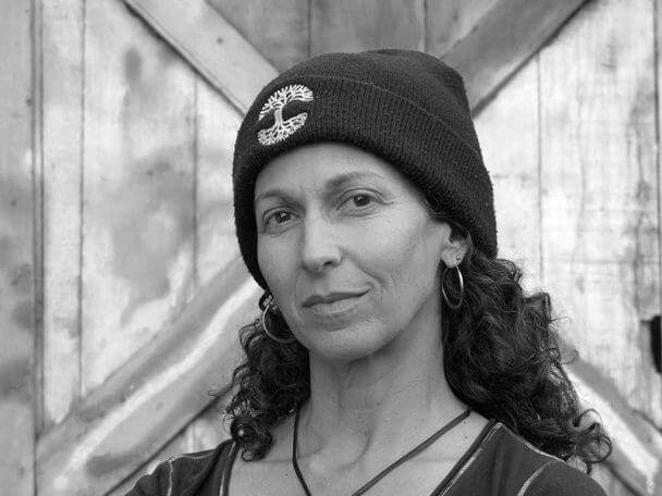 Jane Greenburg looking directly at the camera. She wears a beanie standing in front of a wooden door. Black and white portrait.
