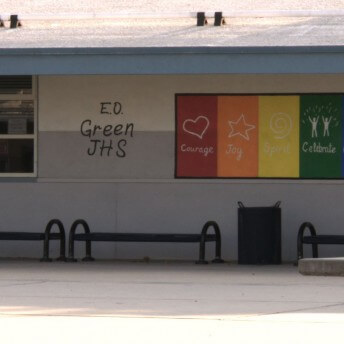 Still from Valentine Road. A building with a window and, text saying, "E.O. Green JHS", and a board displaying a rainbow of colored sections, symbols, and words such as, "courage", "joy", "celebrate", "community", etc.