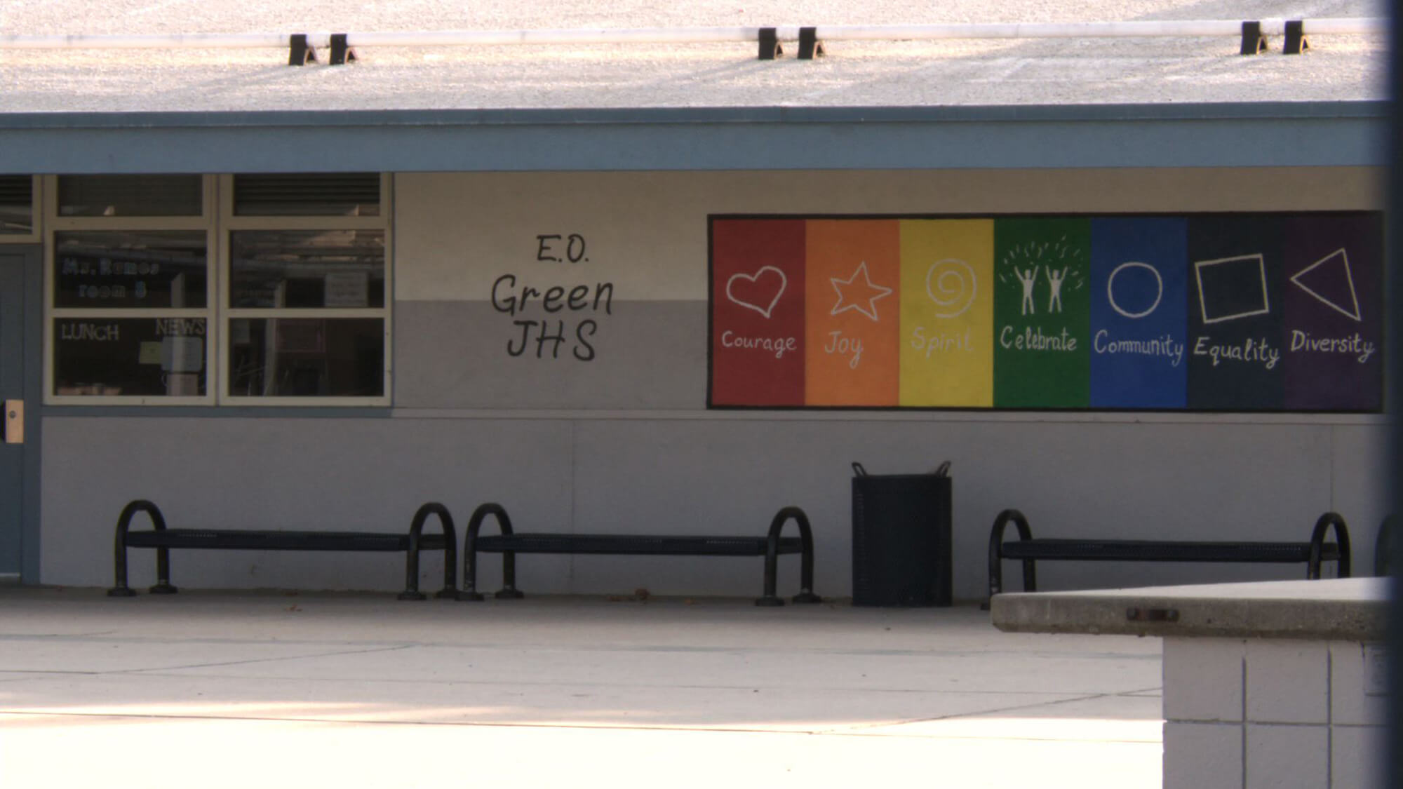Still from Valentine Road. A building with a window and, text saying, "E.O. Green JHS", and a board displaying a rainbow of colored sections, symbols, and words such as, "courage", "joy", "celebrate", "community", etc.