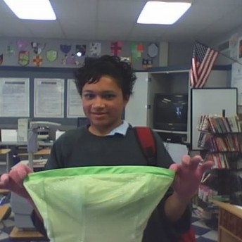 Still from Valentine Road. A young person in a classroom is holding the bodice of a green dress up, smiling at the camera.