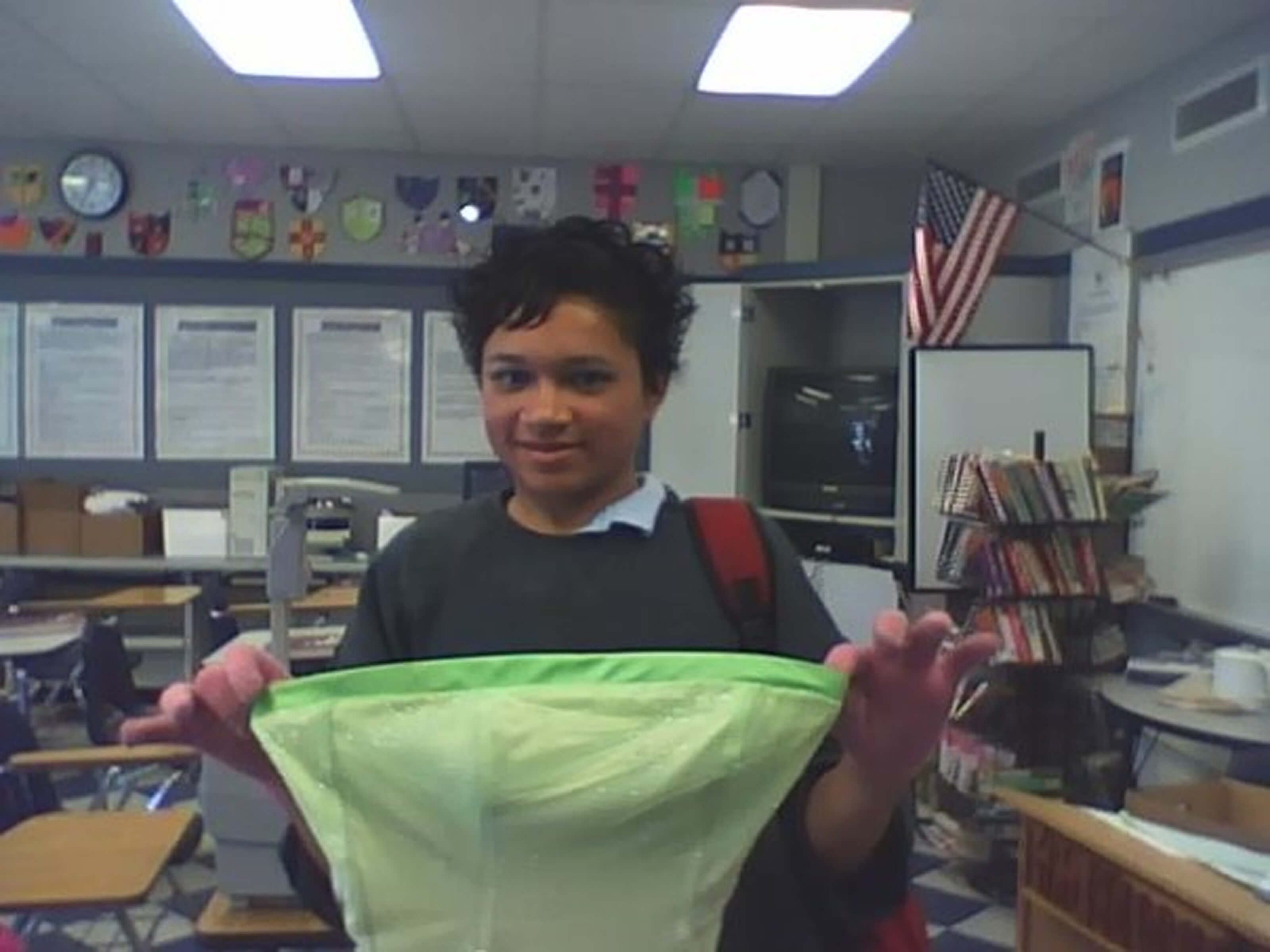 Still from Valentine Road. A young person in a classroom is holding the bodice of a green dress up, smiling at the camera.