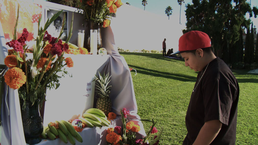 Still from Valentine Road. A young boy stands in front of a memorial for someone. Flowers, candles, and fruit are left behind underneath a portrait of a person.