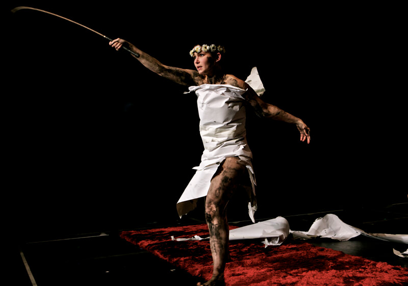 A woman in a white dress with a flower crown is jumping and extending her right arm holding a short sword.