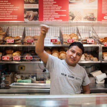 A person behind a restaurant counter pumps their fist in the air. They are standing in front of baskets of bagels organized in bins on the wall.