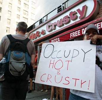 A still from The Hand That Feeds. A person in a white t-shirt and baseball hat holds a sign in front of a storefront. The storefront sign reads, "Hot & Crusty". The sign reads "Occupy Hot & Crusty!"