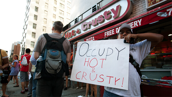 A still from The Hand That Feeds. A person in a white t-shirt and baseball hat holds a sign in front of a storefront. The storefront sign reads, "Hot & Crusty". The sign reads "Occupy Hot & Crusty!"