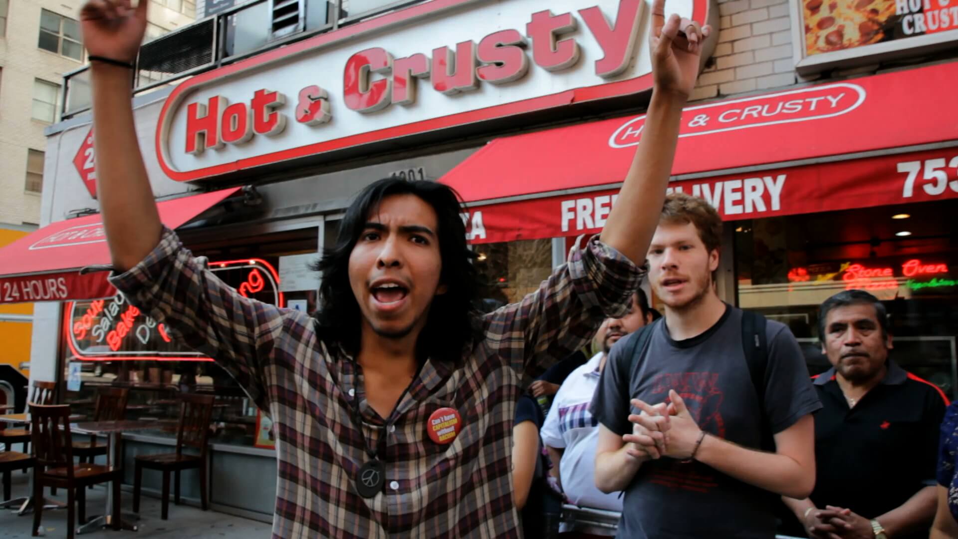 A group of people stands in front of a restaurant. The restaurant sign says "Hot & Crusty". The front-most person in the group holds both of their fists in the air and looks directly at the camera.