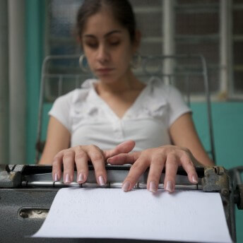 Still from Tocando la Luz. A woman is sitting and touching a machine that has paper with braille coming out of it.