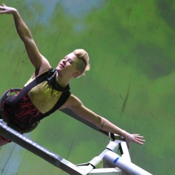 A woman with a mohawk hairstyle is practicing aerial dancing, with her body fully extended, pointed toes, and extended arms.