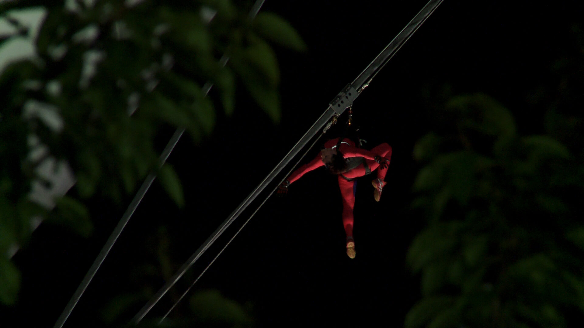 In the night, a human figure wearing a red body suit is hanging from a cable.