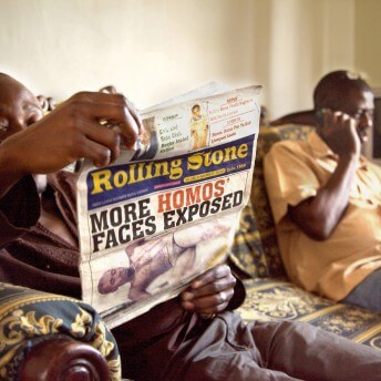 Two men are sitting on a couch, one reads a newspaper, and the other one uses a cellphone.