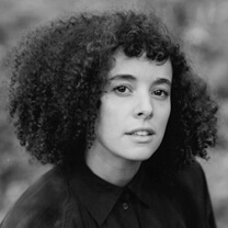 Malika Zouhali Worrall looks directly at the camera. She has medium-length curly hair and is wearing a buttoned shirt. Portrait in black and white.