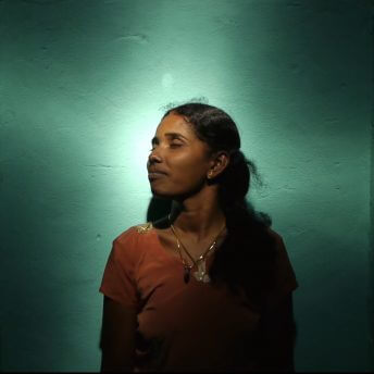 Selvi is looking away from the camera, with eyes closed and smiling. She is standing in front of a green aquamarine wall, with lightning that spotlights her figure.