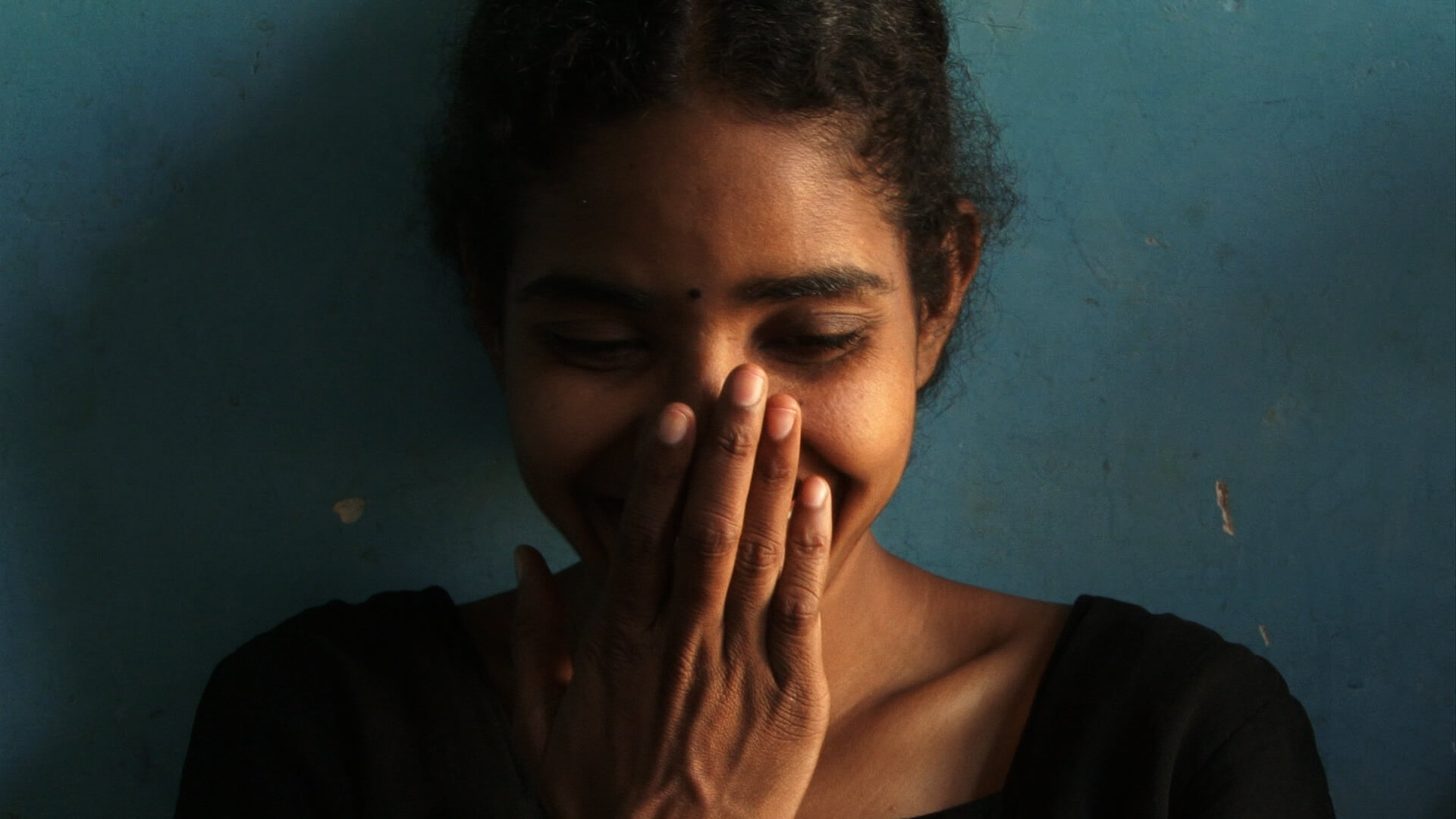 Close up to Selvi's face. She is laughing and placing her hand in front of her face.