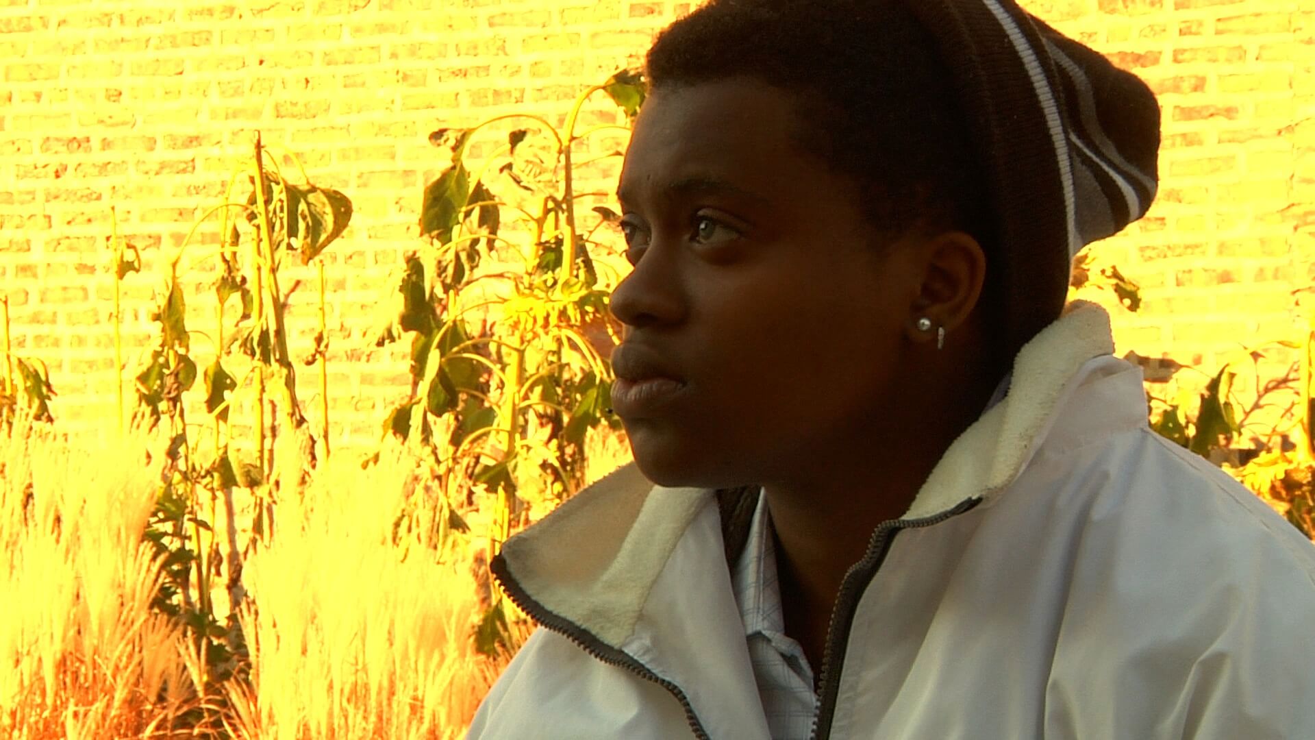 A still from The Homestretch. A close-up shot of a person in a white jacket and maroon beanie outside, in front of a brightly lit wall in the background. The wall appears yellow from the bright sun.