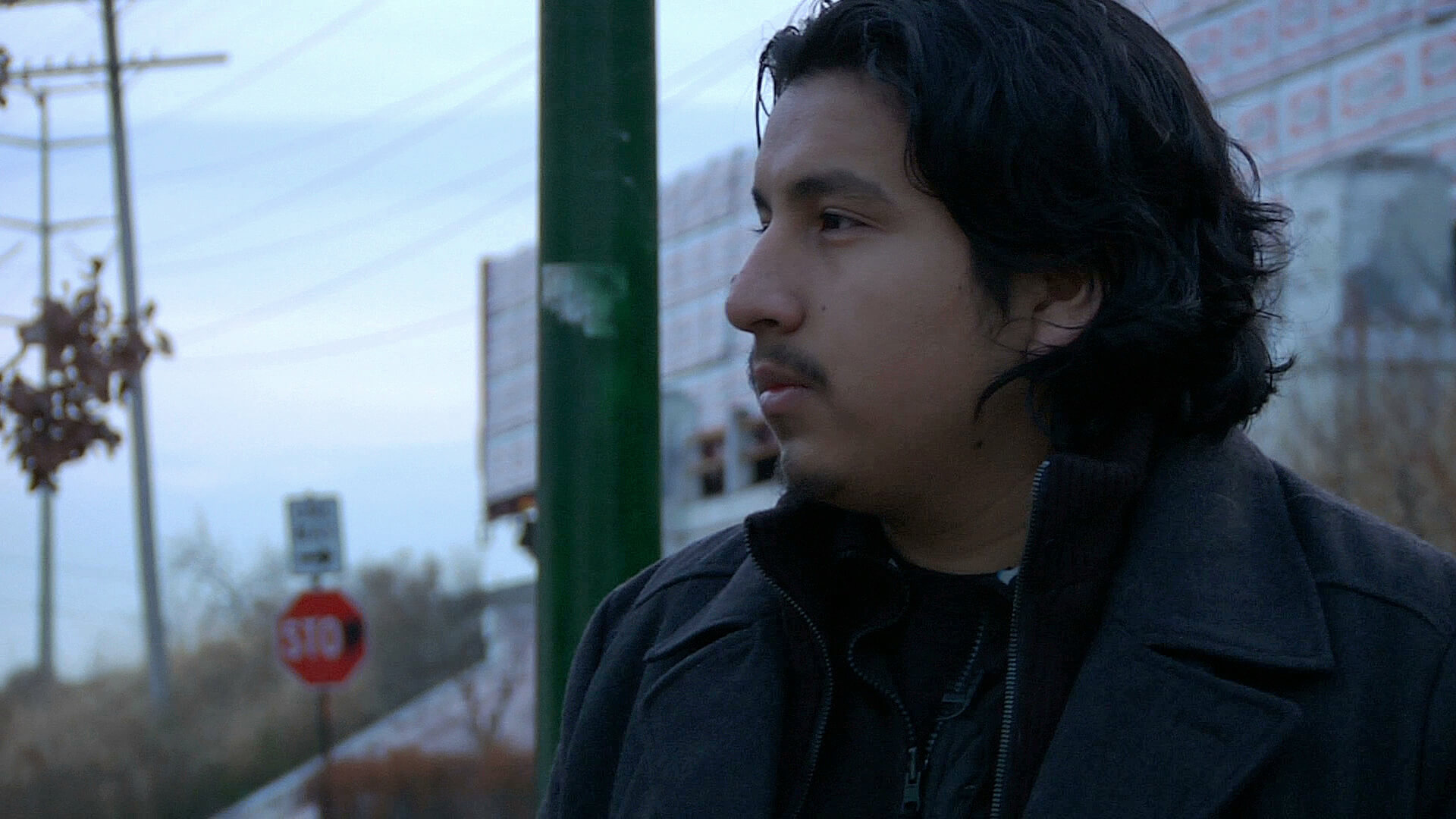 A still from The Homestretch. A close-up shot of a person facing the camera, and looking off to the left of it. They are wearing a black jacket. It is daytime and the sky is cloudy.