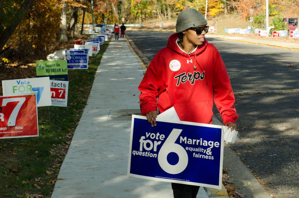 Still from The New Black. A person wearing a hat, sunglasses, and a bright red sweatshirt walks down a sidewalk. They are holding a sign that reads: "Vote for Question 6: Marriage Equality & Fairness".