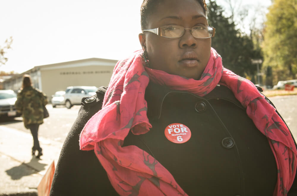 Still from The New Black. A person stands facing the camera and looks directly into the lens. They are wearing a black button-down jacket and a red scarf. They also have a red pin that reads "Vote For Question 6".
