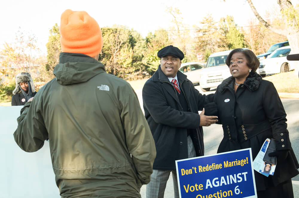 Still from The New Black. Two people stand behind a sign that reads, "Don't redefine marriage! Vote Against Question 6". Another person stands in front of the sign, facing the two people behind it. They are wearing an orange hat and green jacket, their back is to the camera.