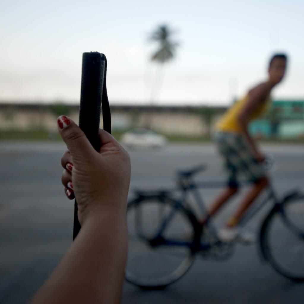 Still from Tocando la Luz. In the foreground, a hand with chipped red nail polish holds the tip of a cane; in the background, an out-of-focus person rides a bike.
