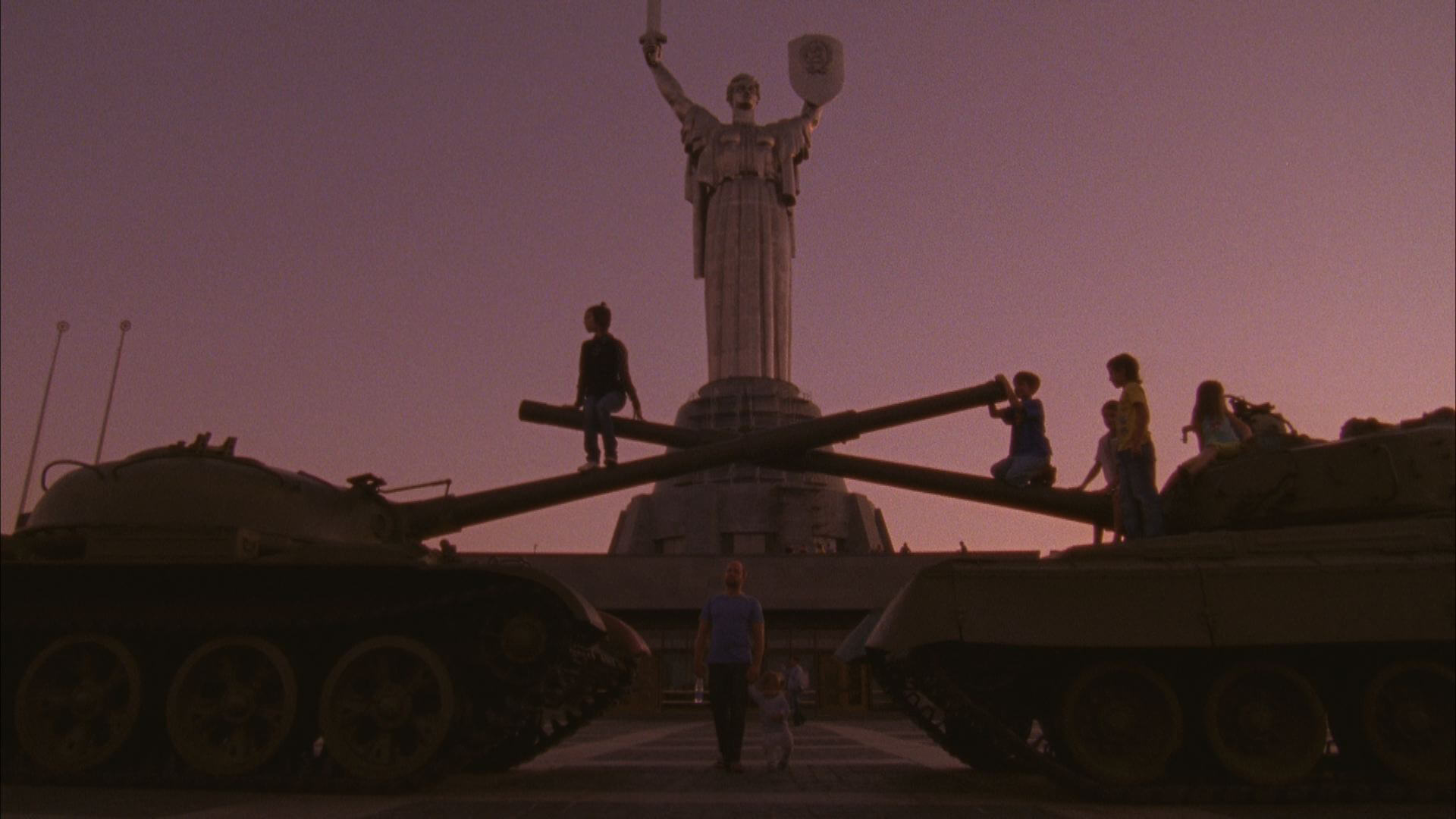 Still from The Vanquishing of the Witch Baba Yaga. Two army tanks face each other with children atop. A large statue stands tall in the background. Color photograph.