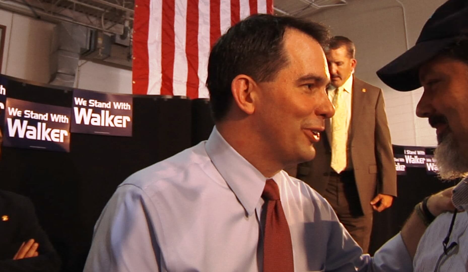A man wearing a shirt and a tie is greeting people out of frame. In the back, there are posters that say "We Stand with Walker."