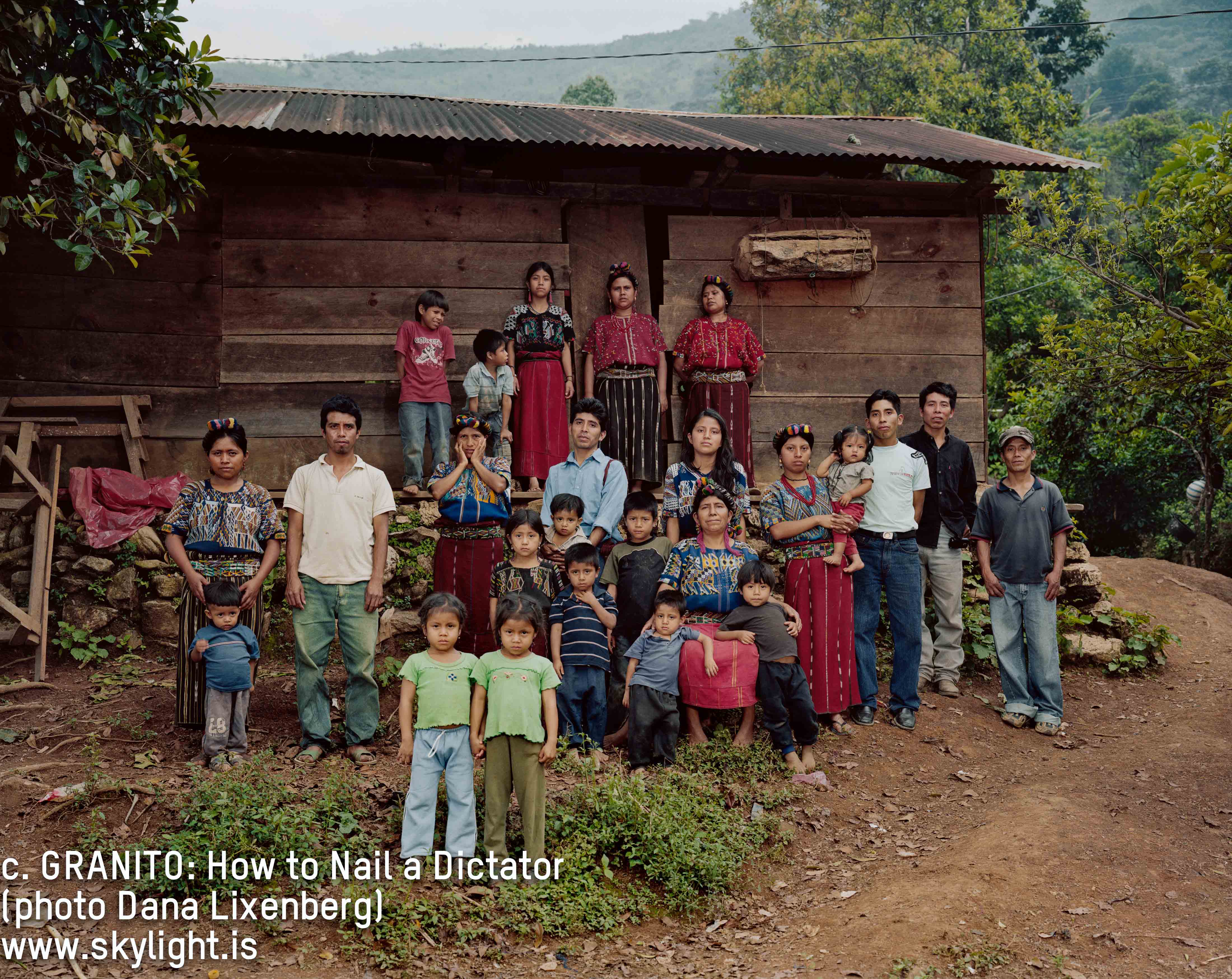 Still from Granito. Outdoor group photo of about 25 people–including, men, women, and children. The women wear patterned traditional Guatemalan indigenous clothing and headbands. All people are staring straight ahead, unsmiling. Photo credit, Dana Lixenberg, is listed on the bottom left of the image.