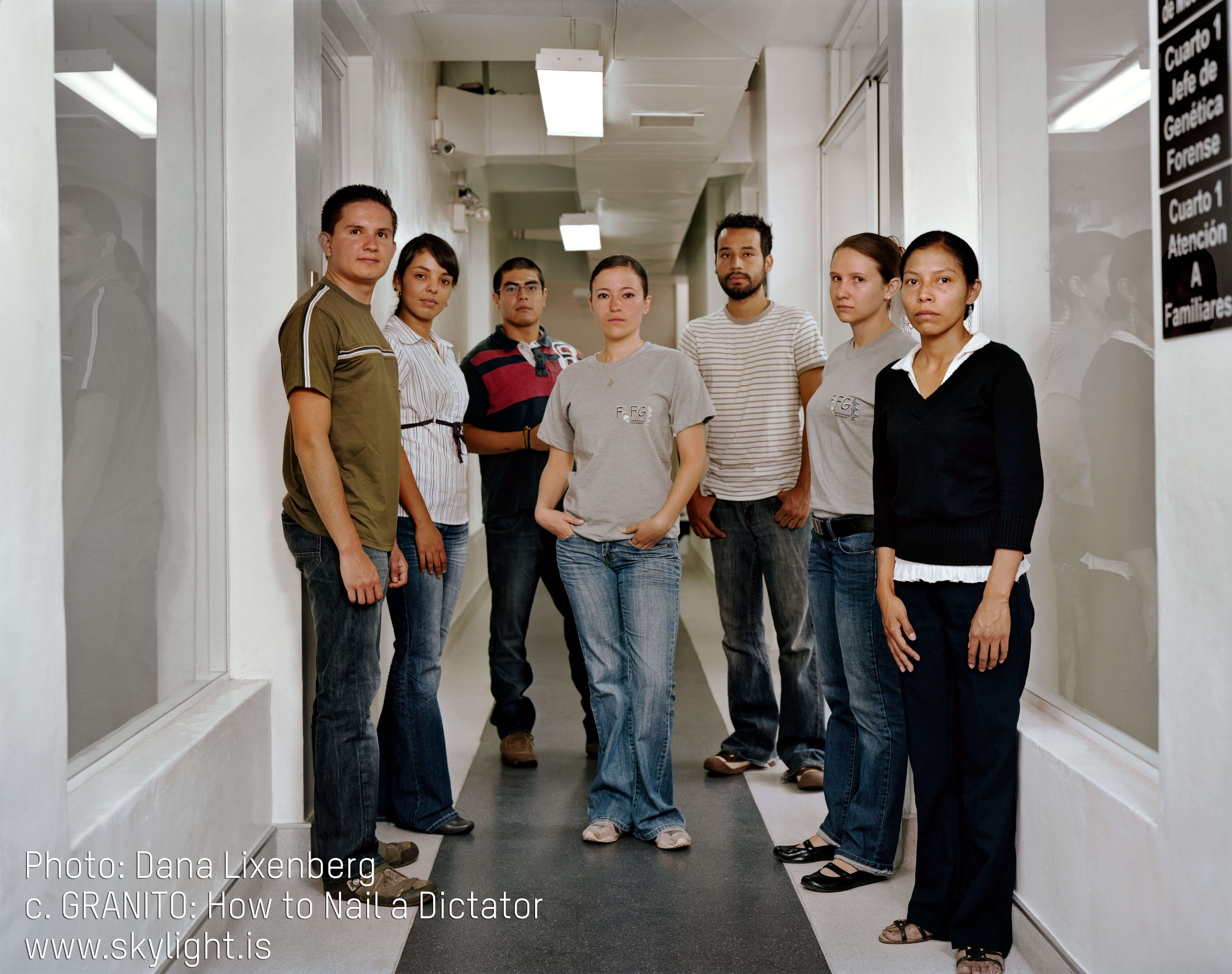 Still from Granito. Group photo of 7 men and women staring straight ahead, unsmiling. The photo is taken in a white hallway, with overhead fluorescenet lights and airducts. An sign on the wall reads: "Cuarto Jefe de Genética Forense" (translation in English: Room of Head of Genetic Forensics). Photo credit is listed on the bottom left of the image.