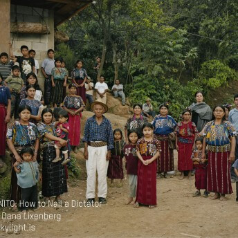 Still from Granito. Outdoor group photo of 30 plus people–mostly women and young children. The women wear patterned indigenous Guatamelan clothing and headbands. All people are staring straight ahead, unsmiling. Director Pamela Yates is seen in the right side of photo, wearing a gray shawl. Photo credit, Dana Lixenberg, is listed on the bottom left of the image.