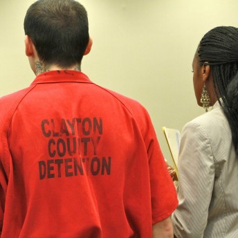 Still from Gideon's Army. A man and woman stand side-by-side and are seen from behind. The man has short hair, black neck tattoos, and is wearing a red jumpsuit with the words "Clayton County Detenion" in black letters on the back. The woman next to him wears her hair in braids, has dangling earrings, and is wearing a blazer and holds a folder with papers.
