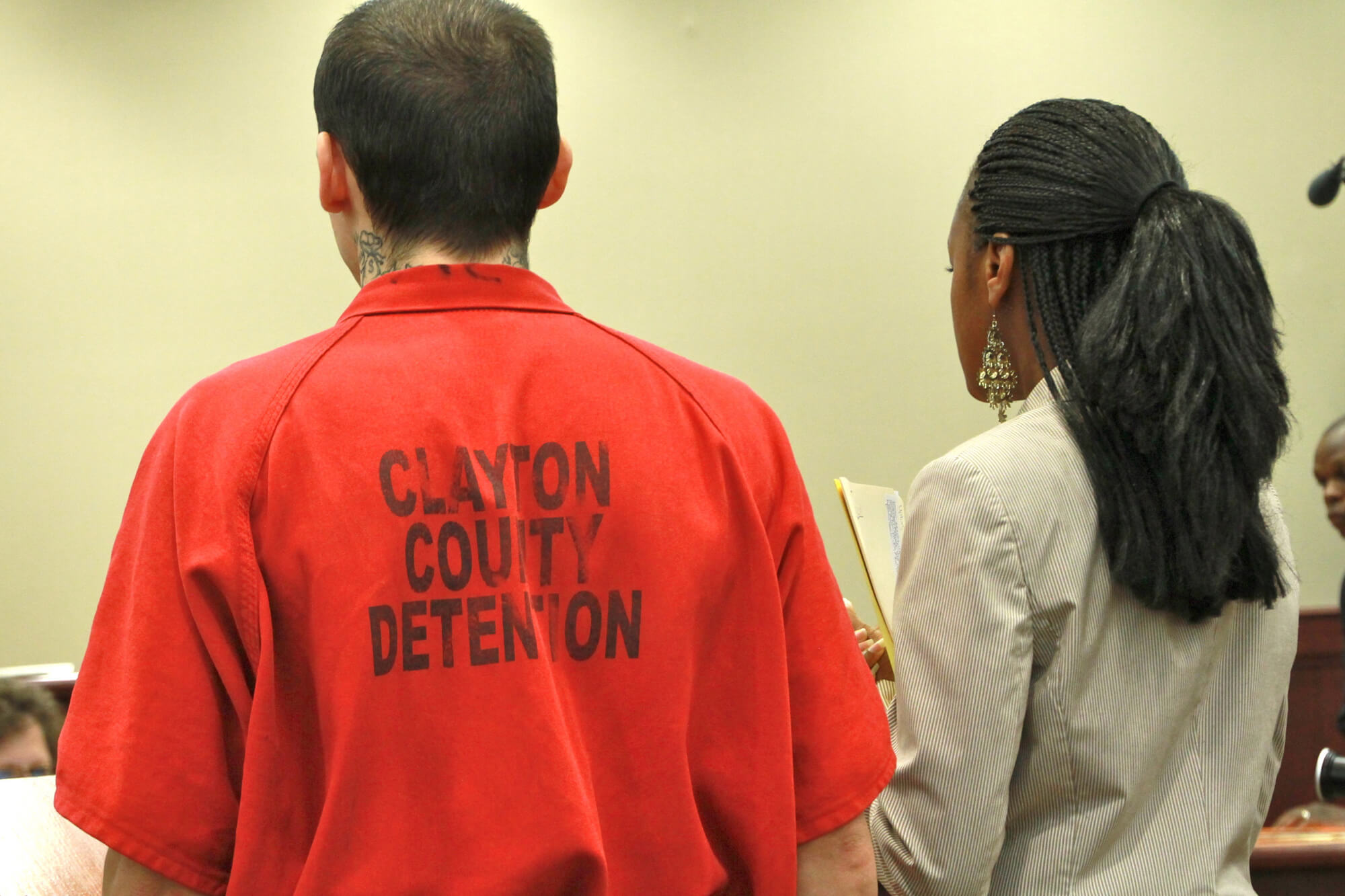 Still from Gideon's Army. A man and woman stand side-by-side and are seen from behind. The man has short hair, black neck tattoos, and is wearing a red jumpsuit with the words "Clayton County Detenion" in black letters on the back. The woman next to him wears her hair in braids, has dangling earrings, and is wearing a blazer and holds a folder with papers.