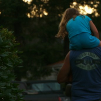 Still from Tough Love. A person is carrying a young girl on their shoulders, and both of them are turning their backs to the camera. There is a pine tree to their left.