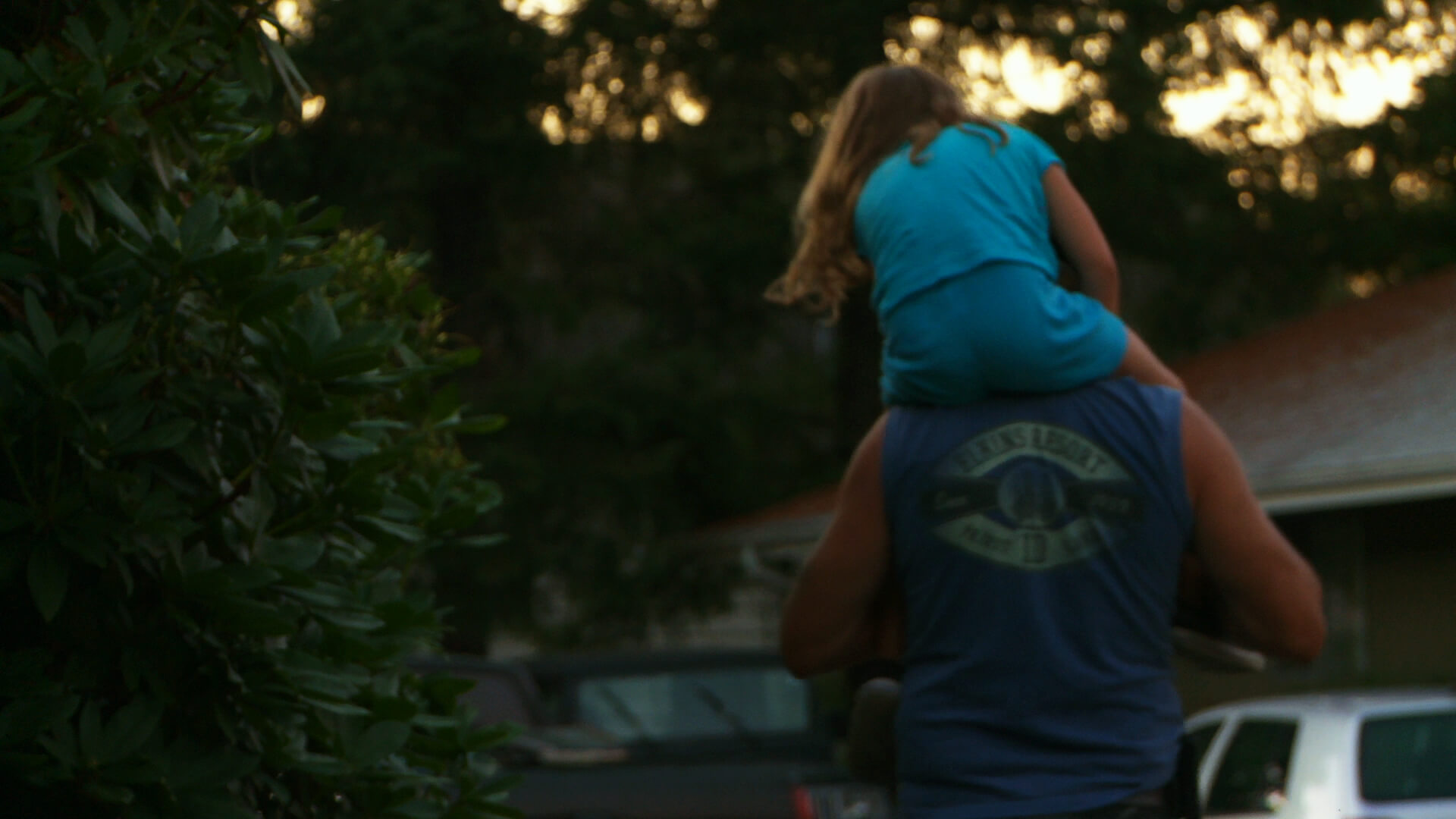 Still from Tough Love. A person is carrying a young girl on their shoulders, and both of them are turning their backs to the camera. There is a pine tree to their left.