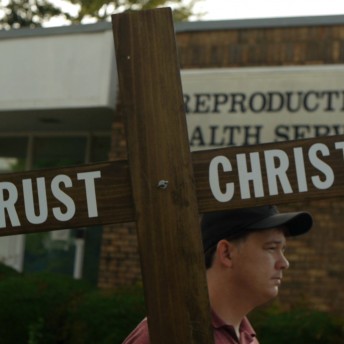 Still from Trapped. A man carrying a wooden cross that says "Trust Christ", he is in front of a Reproductive Health Services clinic.