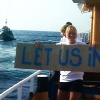 Still from Vessel. A young girl is standing on a boat in the middle of the ocean with a sign that reads, "Let us in!". There is another boat nearby in the water.