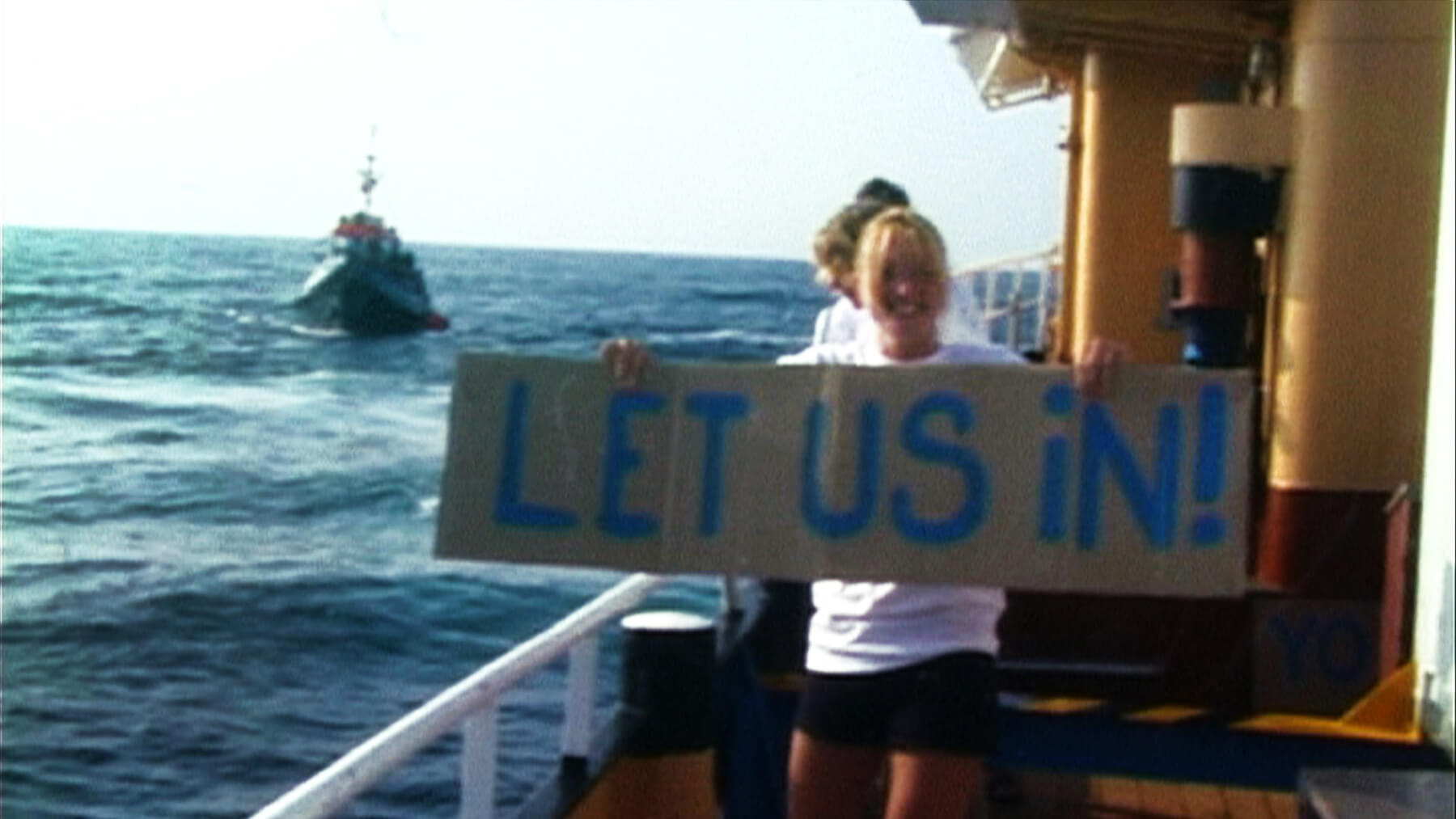 Still from Vessel. A young girl is standing on a boat in the middle of the ocean with a sign that reads, "Let us in!". There is another boat nearby in the water.