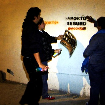 Still from Vessel. Three people are standing by a concrete wall in the dark. The wall is lit by a light, and they had just spray-painted a stencil on the wall.