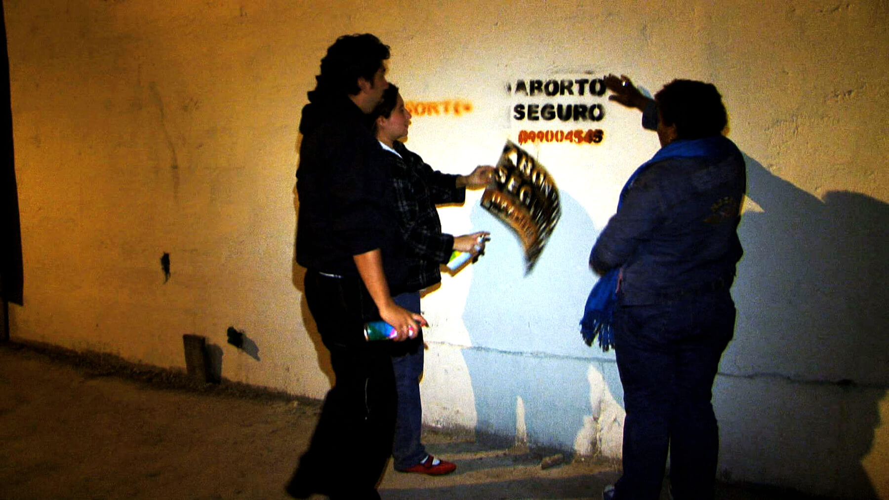 Still from Vessel. Three people are standing by a concrete wall in the dark. The wall is lit by a light, and they had just spray-painted a stencil on the wall.