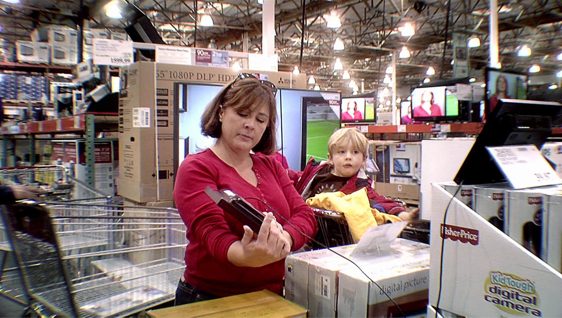 Still from Xmas Without China. A woman stands in an electronics store, holding an electronic with a long cord in one hand. She wears a red top, sunglasses on top of her head, and has short dark hair. A young child sits in a cart behind her. He wears a coat and has blonde hair.