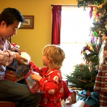 Still from Xmas Without China. A man with dark hair, wearing a striped shirt and jeans, sits and opens a toy with a child. The child, wearing a long sleeve red Santa pajamas, has blonde hair. A young girl with frizzy hair and plaid pajamas stands behind him and next to a decorated Christmas tree.