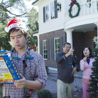 Still from Xmas Without China. A man stands outside holding a small box, with Christmas lights around his neck. He wears a Santa hat and a plaid shirt. An older man and women stand behind him in front of a white and brick home with a Christmas wreath on it.