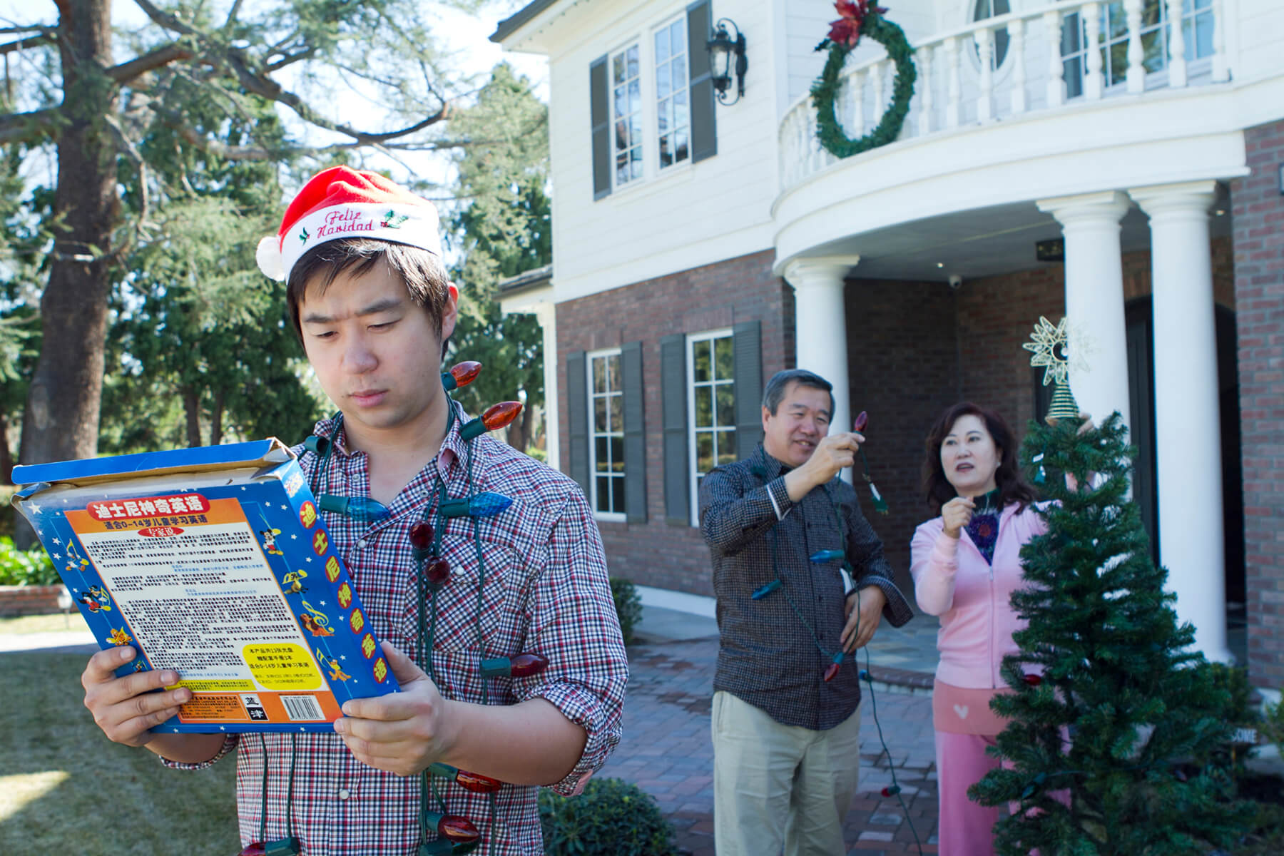 Still from Xmas Without China. A man stands outside holding a small box, with Christmas lights around his neck. He wears a Santa hat and a plaid shirt. An older man and women stand behind him in front of a white and brick home with a Christmas wreath on it.